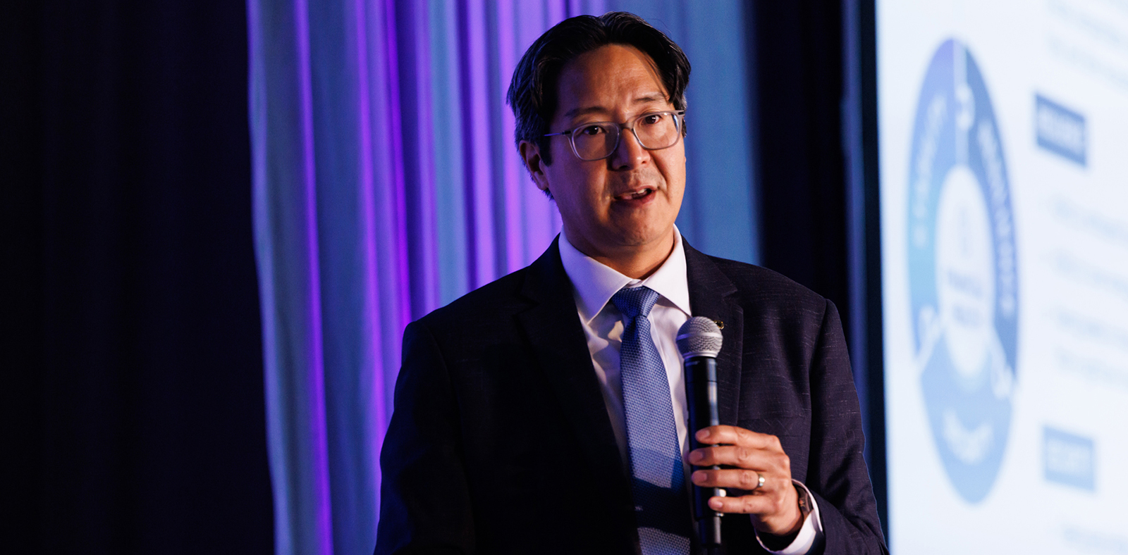 Acting Comptroller Hsu, wearing a dark suit and light blue tie, speaks into a microphone on stage, with purple and blue lighting behind him and a projected slide partially visible in the background.