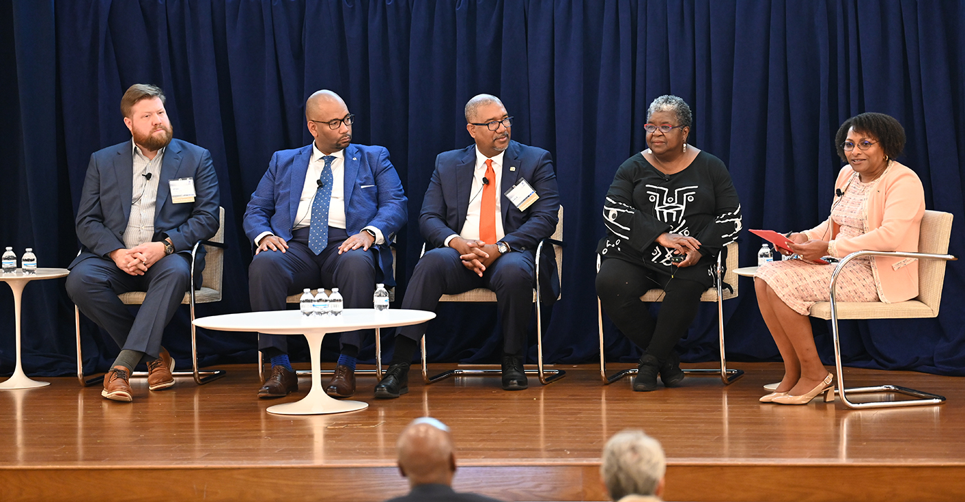 Panelists sit onstage during a discussion at a Project REACh event, with Grovetta Gardineer, the moderator, seated at the far right holding a red folder, against a dark blue backdrop.